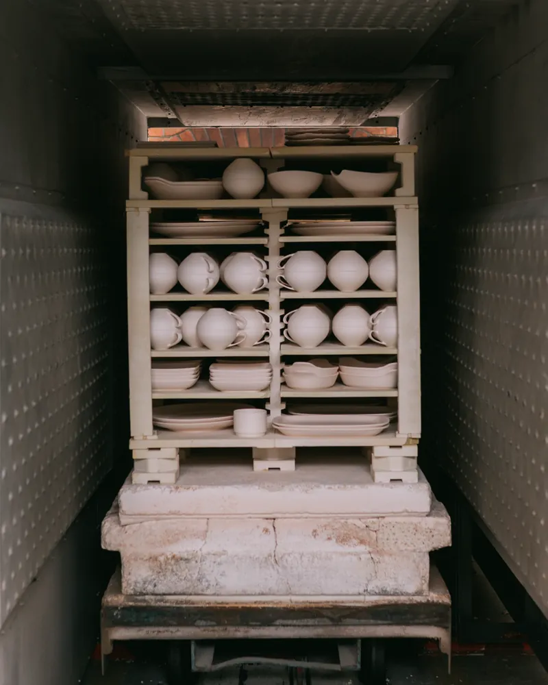 Interior de um forno cerâmico carregado com prateleiras de peças por vidrar: canecas, taças e pratos brancos empilhados sobre placas refratárias